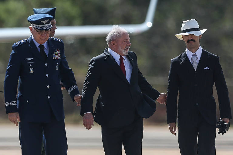O presidente Luiz Inácio Lula da Silva participa de Cerimônia alusiva ao sesquicentenário de Alberto Santos Dumont na Base Aérea de Brasília. Ele caminha junto a outros homens de terno. Ao fundo, vê-se parte de um avião