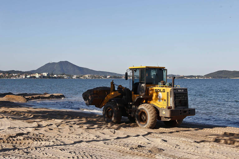 Uma escavadeira amarela atravessa uma praia. A areia é batida e a água, azul, não tem ondas. Ao fundo, pequenos morros. O céu é azul