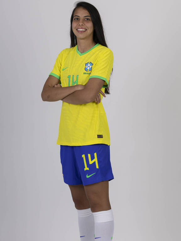 A zagueira Lauren, da seleção brasileira de futebol, posa para foto com os braços cruzados; ela usa camisa amarela, calção azul e meiões brancos