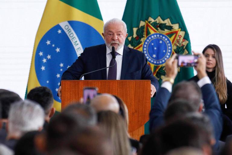 Lula, um homem branco de cabelo e barba branca vestido de terno, fala para plateia. Ao fundo há o brasão e a bandeira do Brasil.