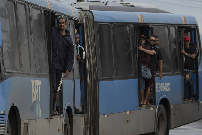 Lateral de um ônibus azul com pessoas em pé, com parte do corpo para fora. Há ao menos quatro homens em pé nas portas do veículo
