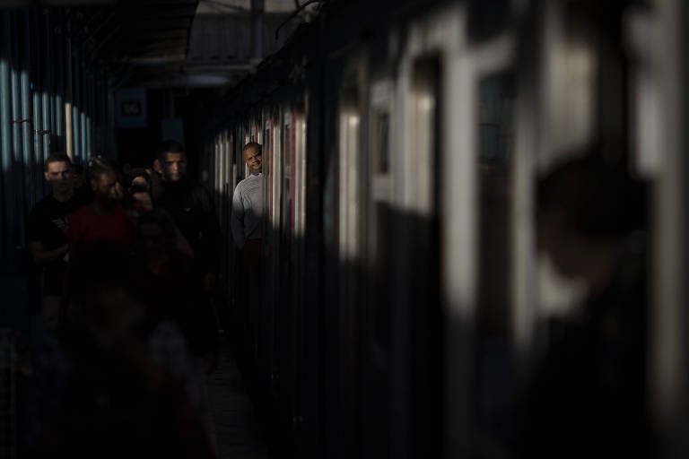 Homem dentro de vagão de trem olha para frente. Ao seu lado, pessoas caminham pela estação. A luz do fim da tarde cobre parte do trem