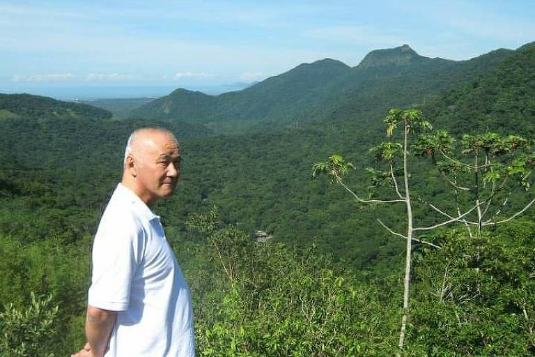 homem idoso de origem japonesa com cabelos brancos ralos vestindo camisa branca em frente a paisagem de montanha e mata