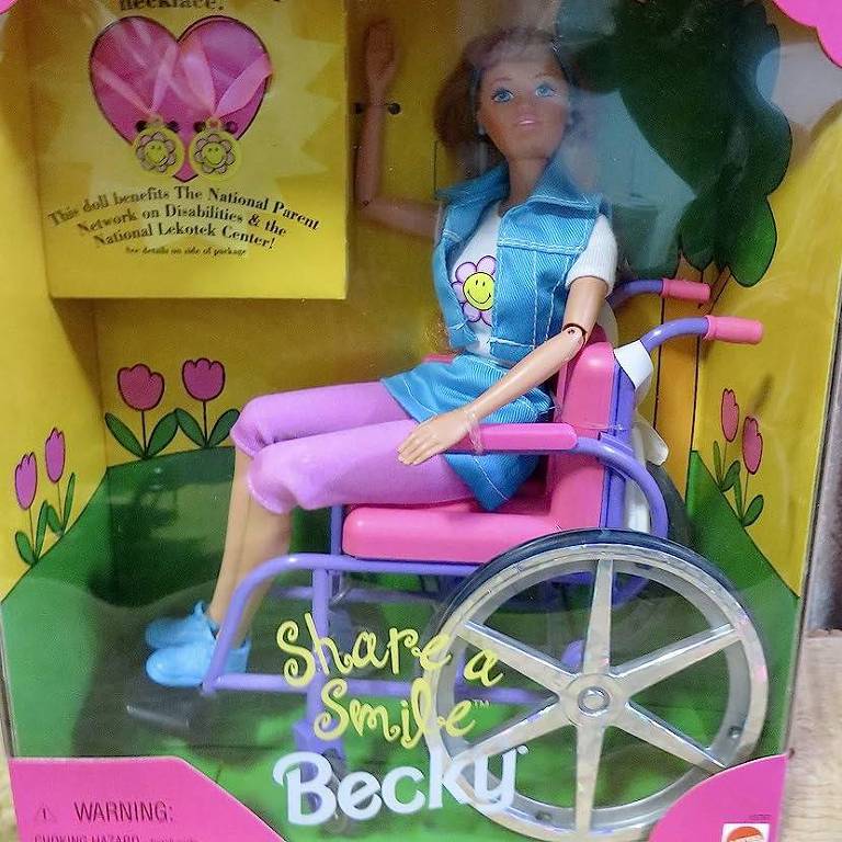 Uma boneca da Barbie, com bermuda cor-de-rosa, sapatos e blusa azuis, sentada em uma cadeira de rodas. O brinquedo está dentro da caixa original