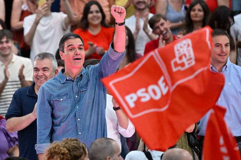 Homem branco de meia idade, cabelo curto e camisa cinza com punho esquerdo erguido com multidão e bandeira vermelha do PSOE ao fundo