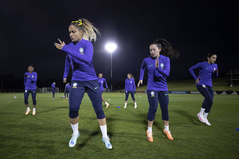 Mulheres vestidas com roupa esportiva azul treinam futebol em gramado à noite