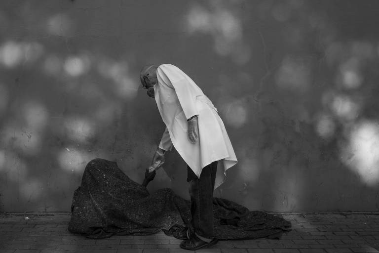 Foto em preto e branco com tonalidades mais escuras mostra uma pessoa de jaleco branco em pé entregando um pão a uma pessoa deitada no chão