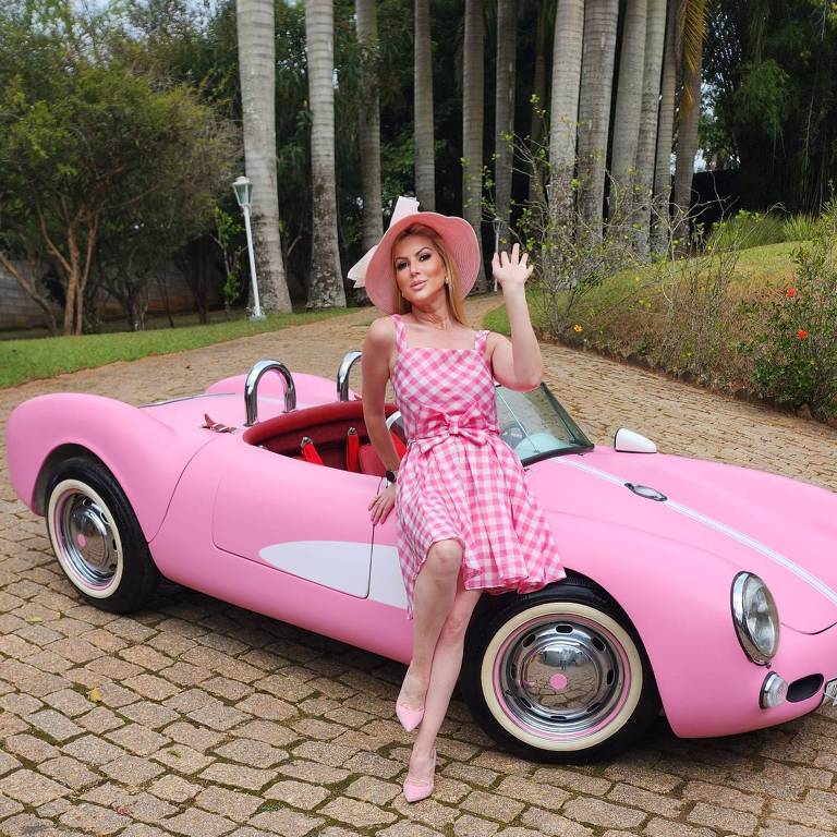 a gerente administrativa Barbie Tavares, com chapéu e vestido rosa, encostada na porta da réplica de um Porsche Spyder rosa
