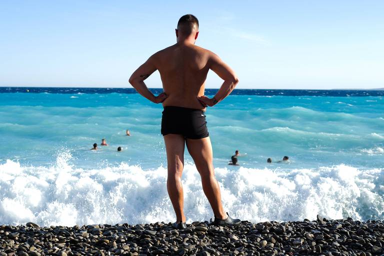 Homem de pé, de costas, observa pessoas tomando banho no mar