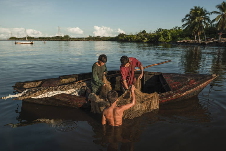 Dois jovens estão em cima de um barco, enquanto um adulto, dentro da água, os ajuda com uma rede de pesca