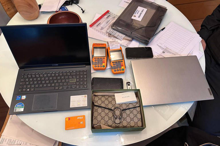 Imagem mostra mesa cheia de produtos como computadores, celulares, pen drive, bolsa, máquinas de cartão e documentos.