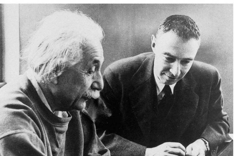 Sentados lado a lado, Robert Oppenheimer e Albert Einstein olham para papel numa mesa à sua frente