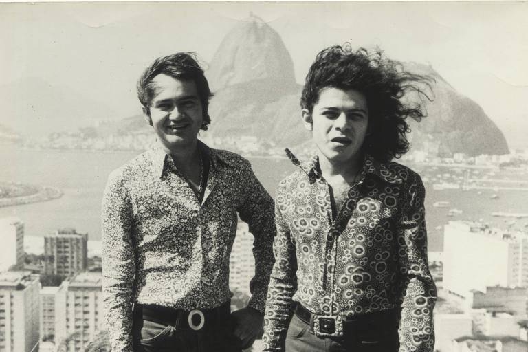 Fotografia em preto e branco de Óscar Ramos e Luciano Figueiredo. Ambos usam calça, cinto com fivela e camisa estampada. Ao fundo, prédios e o corcovado.