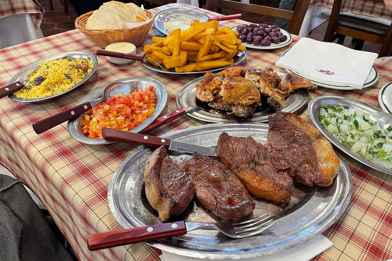 A foto mostra uma mesa com diversas baixelas de inox com farofa, tomate picado, polenta frita, galeto e quatro fatias de picanha