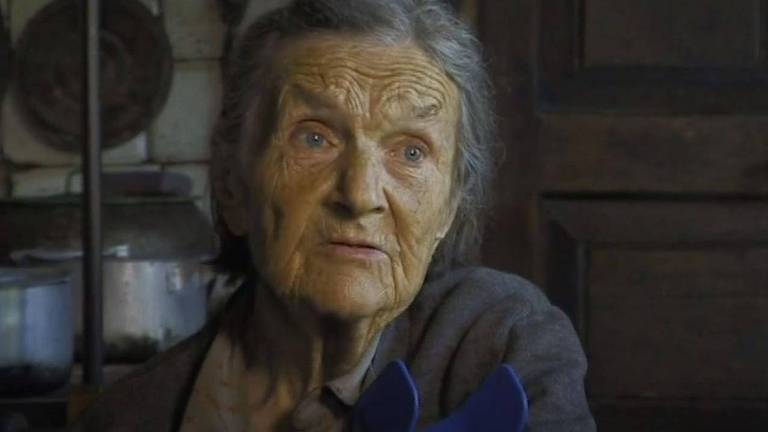 Retrato de Maria, uma mulher branca, idosa, com cabelos grisalhos; ela veste blusa cinza