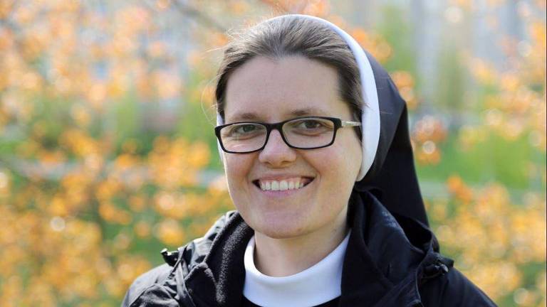 Retrato da freira Klara, uma mulher branca com cabelos castanhos; ela sorri e veste um hábito de freira nas cores preto e branco, além de óculos de grau com armação preta