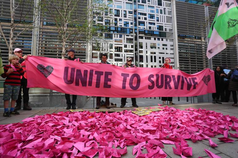 Pessoas seguram cartaz escrito 'unite to survive' (se unam para sobreviver) cor de rosa, diante de muitos barquinhos de papel, também cor de rosa, dispostos em uma calçada