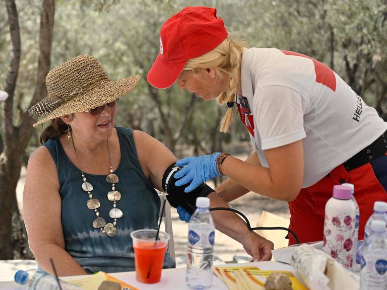 Um agente de saúde da Cruz Vermelha, usando uniforme vermelho e branco, se inclina ao lado de uma mulher usando chapéu e óculos de sol para aferir a pressão sanguínea dela