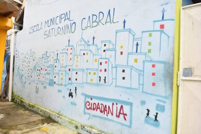 Foto mostra muro da escola com desenhos de prédios feitos em grafite em um muro branco. A palavra "cidadania" está escrita na parede