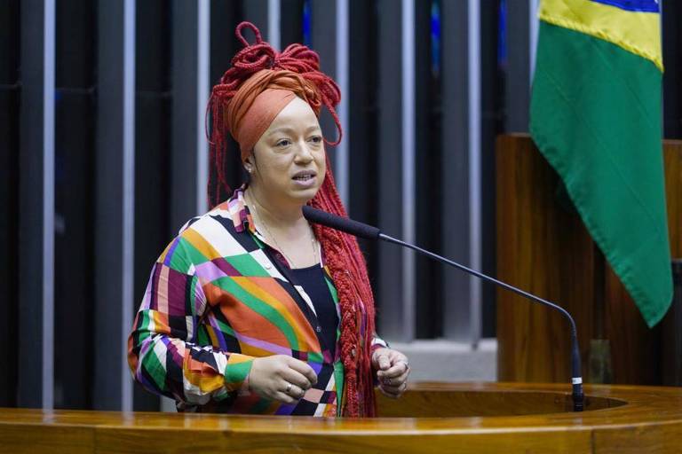 Imagem de uma mulher discursando em um púlpito. Ela está vestindo uma blusa colorida e tem os cabelos trançados e vermelhos. Ao fundo, há uma bandeira do Brasil.