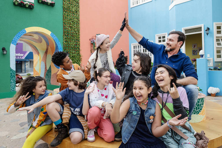 Diversas crianças e um homem adulto estão em um banco de praça fazendo algazarras e sorrindo; elas usam roupas coloridas e ao fundo há várias casas também com cores marcantes