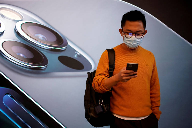 Homem de máscara mexe em smartphone em frente à publicidade de iPhone em loja da Apple, em Shangai, na China. Ele veste agasalho laranja e calças pretas.