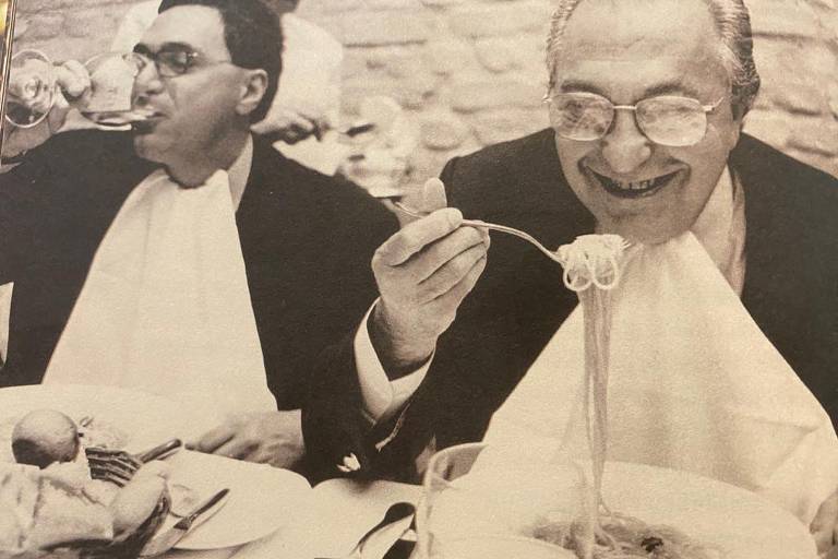 Dois homens sentados à mesa; um deles, mais jovem, bebe um gole de vinho, e o outro, um senhor, dá uma garfada num prato spaghetti, sorrindo