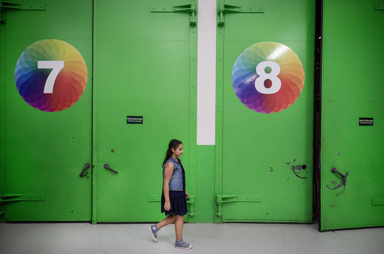 Uma garota de roupas e tênis azuis passa em frente a duas enormes portas de ferro pintadas de verde claro onde se lê os números 7 e 8