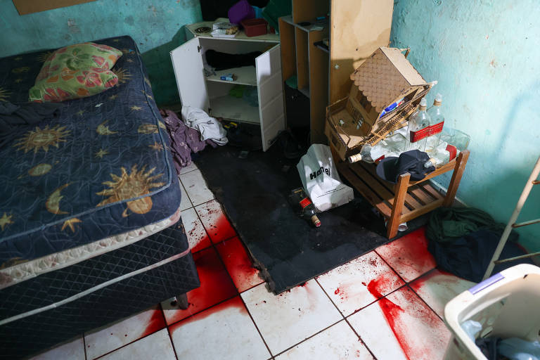 A imagem mostra um quarto desordenado com um colchão em uma cama， um tapete preto no chão e manchas de sangue espalhadas pelo piso. Há um armário branco parcialmente aberto， uma cadeira de madeira com objetos em cima e lixo espalhado pelo chão. As paredes são de uma cor verde desbotada.