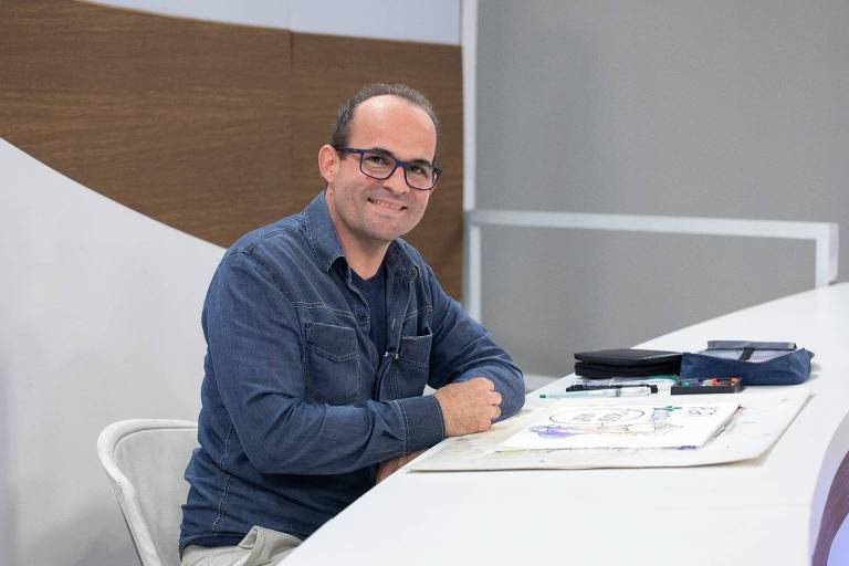 Ilustrador Luciano Veronezi, durante participação no programa Roda Viva. Luciano usa óculos de grau, camisa azul, e está apoiado na mesa do cenário do programa Roda Viva.