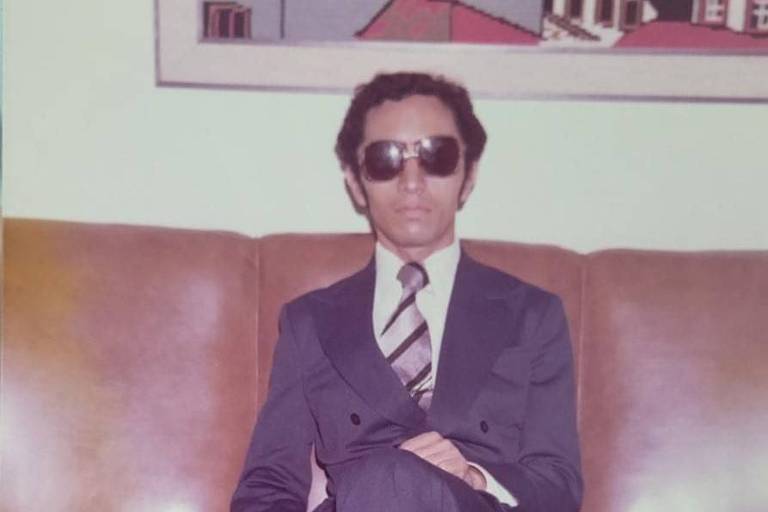 foto antiga, manoel usa óculos escuros, terno escuro, está sentado em um sofá, tem os cabelos pretos penteados de lado