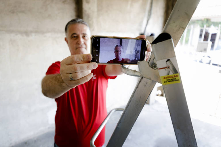 A imagem mostra Ricardo, um homem branco de cabelos grisalhos, segundo um celular, que está apoiado em uma escada. Ele veste uma camisa vermelha