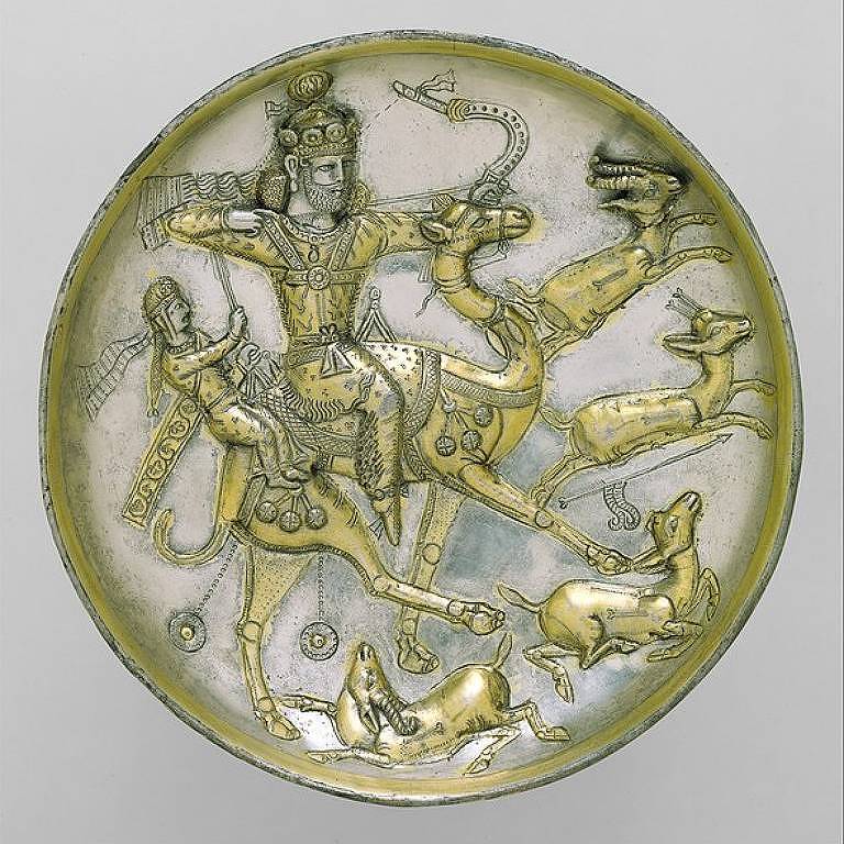 Círculo dourado com figuras esculpidas na parte de dentro, como um homem andando de camelo e atirando uma flecha