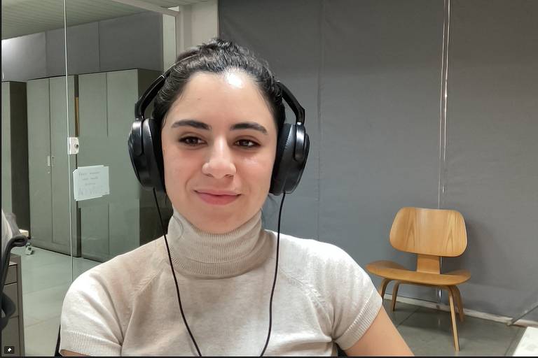 TV Folha reporter Giovanna Stael, without the Maybelline filter.  Giovanna is a white woman, with straight black hair tied in a bun.  She wears a cream turtleneck and wears a black headset.