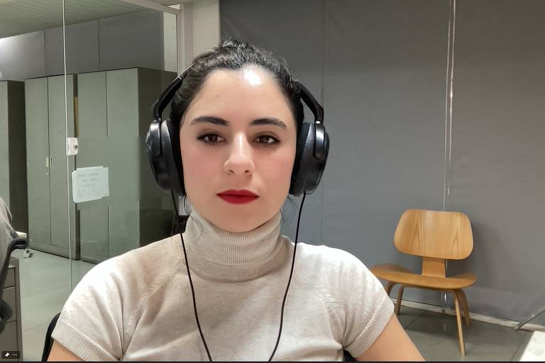 TV Folha reporter Giovanna Stael, with the Maybelline filter -- red lipstick and light eyeshadow.  Giovanna is a white woman, with straight black hair tied in a bun.  She wears a cream turtleneck and wears a black headset.