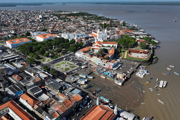 foto aérea mostra Belém, com prédios, áreas arborizadas e igrejas à esquerda, e embarcações na água, do lado direito