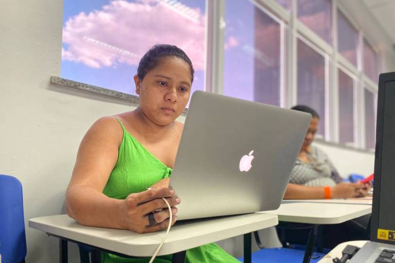 Mulher com traços indígenas olha para um laptop usando um vestido verde sentada numa carteira escolas com uma janela ao fundo