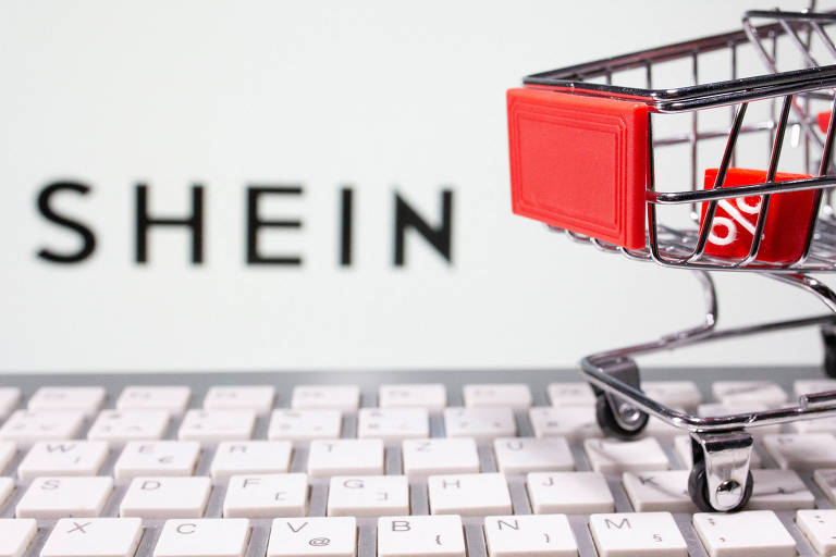 Imagem mostra um teclado e um carrinho de compras em miniatura em frente ao logo da Shein, ligeiramente desfocado
