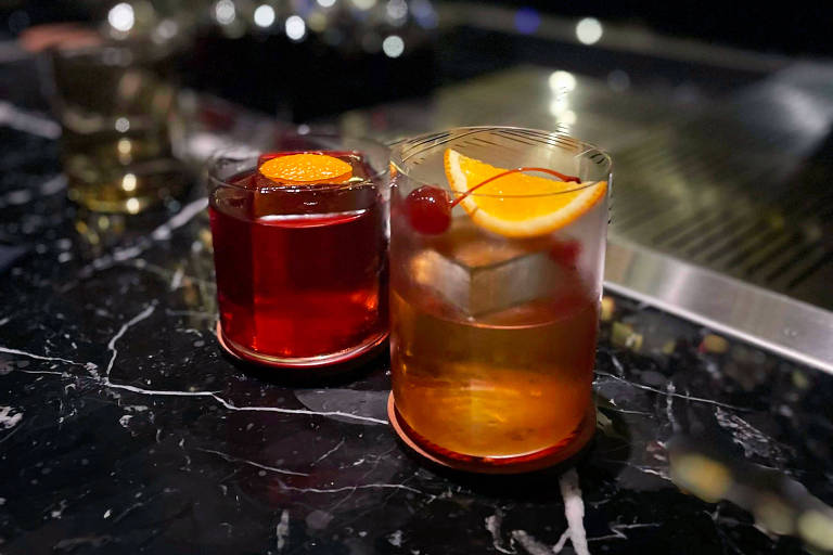 A foto mostra dois copos baixos contendo drinques: o primeiro, com Negroni, traz um líquido avermelhado e o segundo, um Old Fashioned, apresenta um líquido mais alaranjado e vem guarnecido com cereja e uma fatia de laranja.
