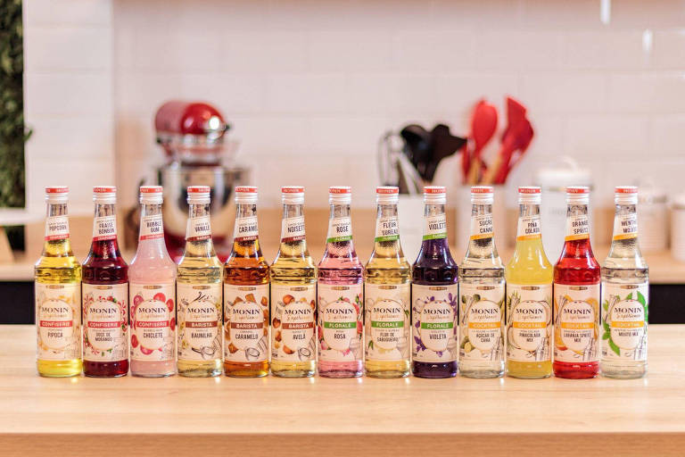 A foto mostra 13 garrafas alinhadas lado a lado com xaropes variados e de diversas cores da marca francesa Monin.