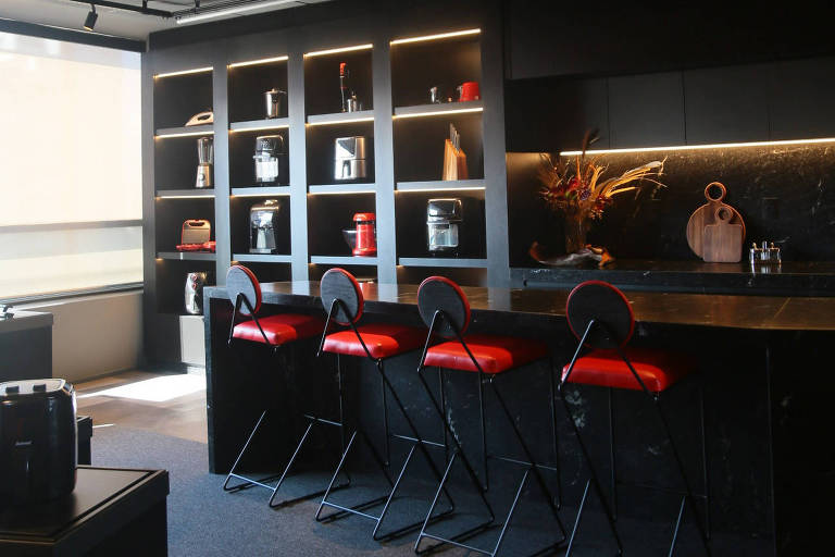 A foto mostra uma cozinha-show pintada de preto com uma bancada e quatro bancos com assento vermelho. Ao fundo, uma parede com nichos ocupados por eletrodomésticos.