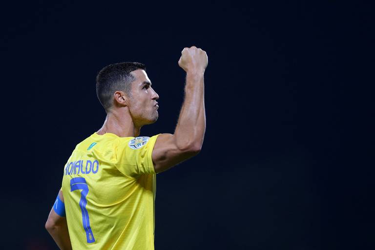 Cristiano Ronaldo aparece celebrando um gol com o punho direito em riste. Ele veste a camisa 7 do Al Nassr. Seu uniforme é amarelo.