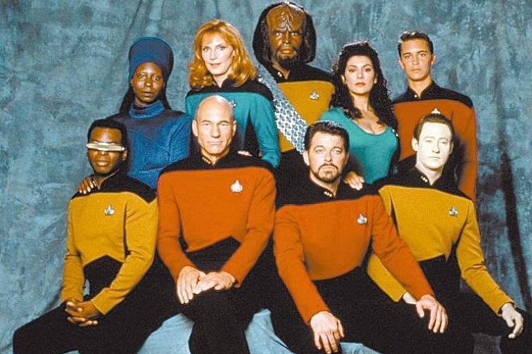 Nove pessoas estão na foto, uniformizados com roupas coloridas da série. Há quatro mulheres, quatro homens e um klingon (de pé, ao centro, na última fileira)