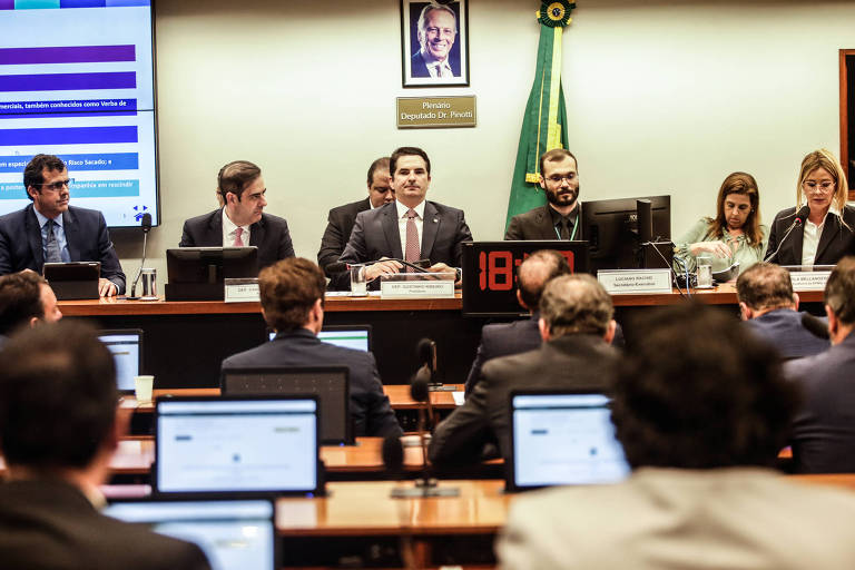 Plenário com laptops à vista e homens de terno diante de bandeira do brasil 