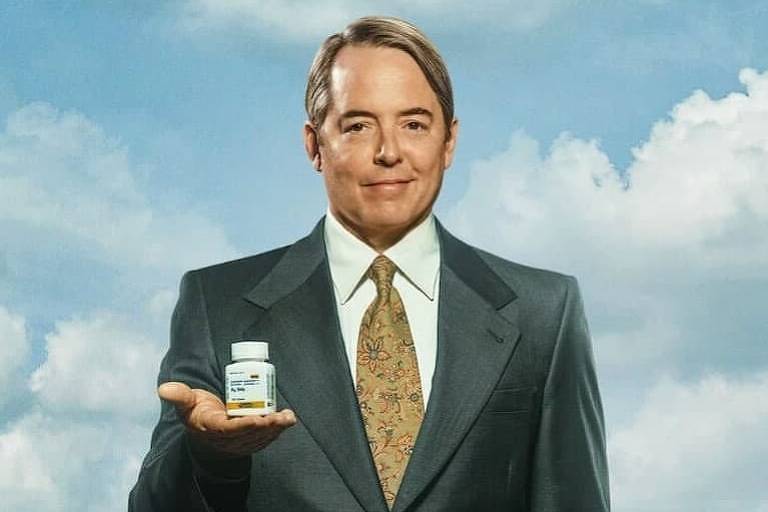 Homem de terno branco e de meia idade sorri para a câmera enquanto segura um frasco de comprimidos; atrás dele se vê pílulas e um céu azul