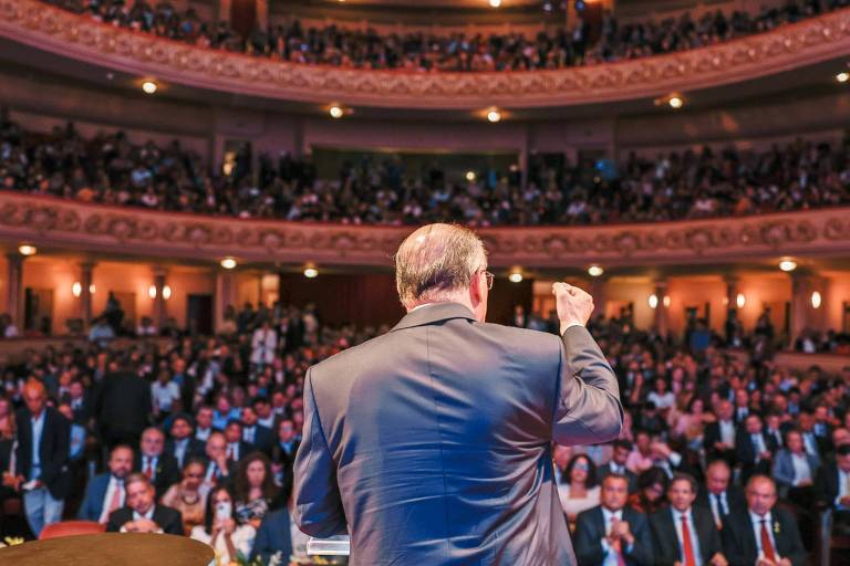 Geraldo Alckmin aparece de costas falando em um púlpito. Ao fundo, está a plateia em um teatro.