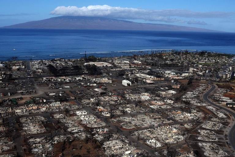 Vista aérea da cidade destruída, com prédios e casas completamente queimados, com cinzas no lugar das quadras; ao fundo é possível ver o mar azul e um vulcão