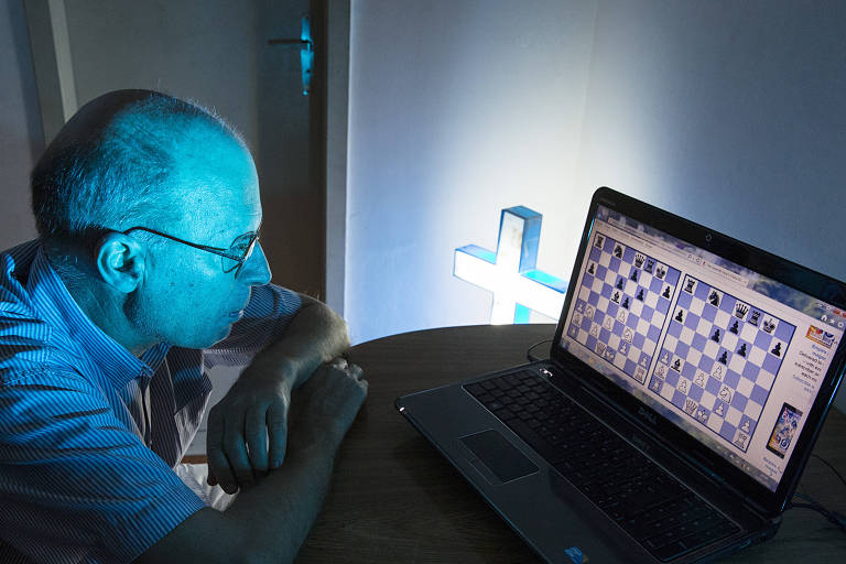 Homem calvo diante de um computador; ao fundo, um crucifixo ilumina o ambiente