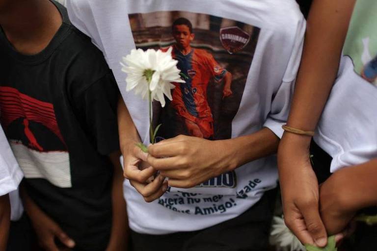 Três adolescentes em enterro, com flores na mão e um deles com camisa estampando foto de Thiago Menezes Flausino