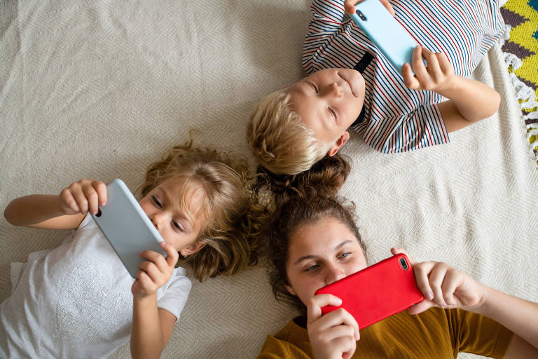 Três crianças deitadas no chão mexem em seis celulares